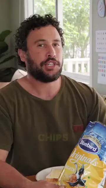 A man at his dining table holding up a bag of chips beside a plate of buttered bread